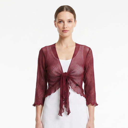 A woman stands against a plain white studio background wearing a sheer burgundy lace shrug with three-quarter sleeves and a front tie, over a white midi dress and white sneakers.