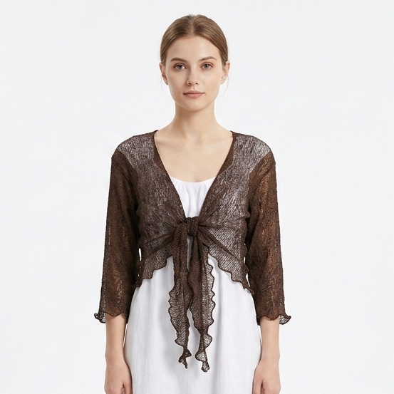 A woman stands against a plain white studio background wearing a sheer dark brown lace shrug or cardigan over a white midi dress and white sneakers.
