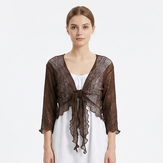 A woman stands against a plain white studio background wearing a sheer dark brown lace shrug or cardigan over a white midi dress and white sneakers.